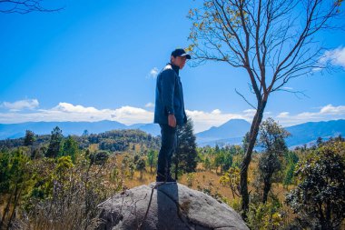 Siyah elbiseli ve şapkalı, kuru ağaçlı bir kayanın üzerinde duran genç adam.