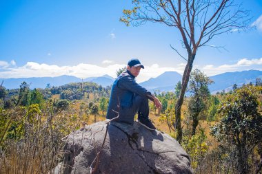 Genç adam güneşin altında siyah giysiler içinde oturuyor ve manzara çok güzel.