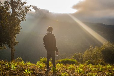 Genç adam siyah giysilerle parlayan güneşe bakıyor.