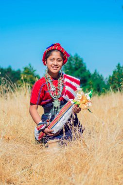 Kuru çimenlerin arasında oturan bir kadın elinde çiçeklerle arka planda ormanla