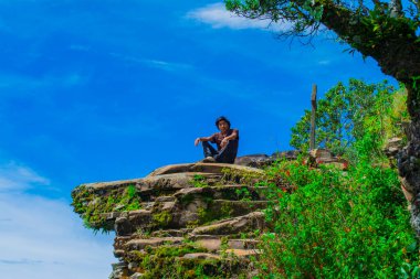 Arkasında haç ve yeşil ağaçlarla kayaların üzerinde oturan genç adam.