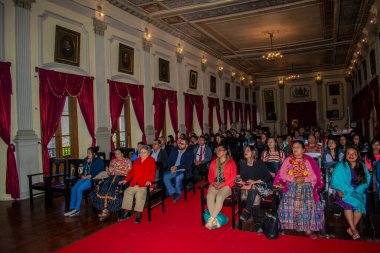 İnsanlar büyük salonda siyah sandalyelerde oturuyorlar.