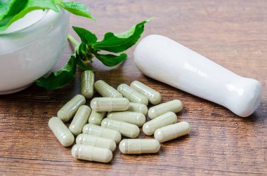 Kapsüllerde bitkisel ilaç yığını ve ahşap arka planda yeşil yapraklı seramik havan..