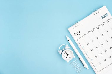 Temmuz 2026, 2026 yılı için aylık masa takvimi ve mavi arkaplanlı ataçlı çalar saat. Kopyalama alanı olan konum.