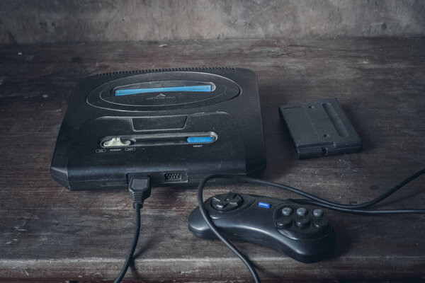 Старая игровая приставка в черном, в пыли на деревянном столе, рядом с джойстиком и картриджем.