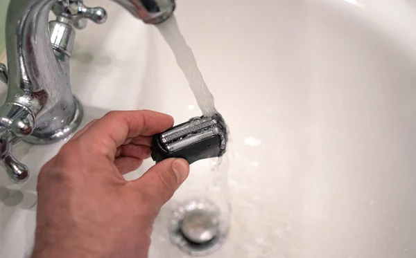 Washing the blades from an electric razor under running water in the sink. A man\'s hand with a plastic razor head.