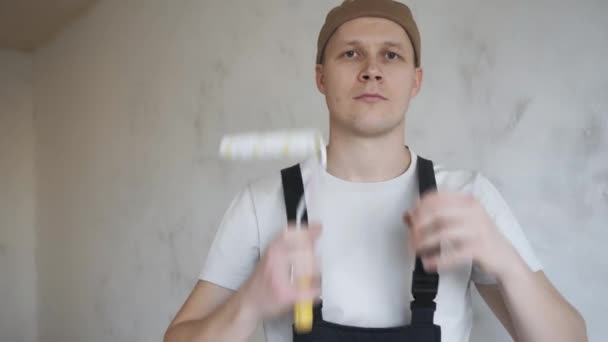 Un hombre en un mono de construcción felizmente muestra un rodillo de pintura a la cámara — Vídeos de Stock