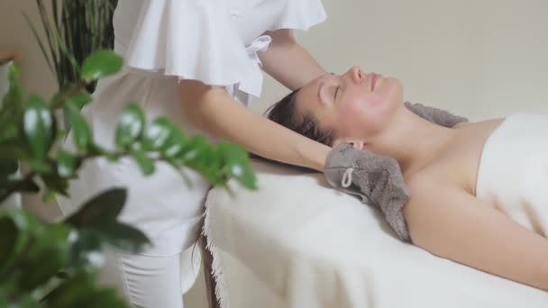 Beautiful girl lies in the spa salon and the master wipes her with special glove — Stock Video