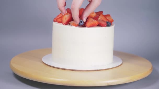 En ung kvinnas händer dekorerar en vit tårta med bär på en trälåda. — Stockvideo