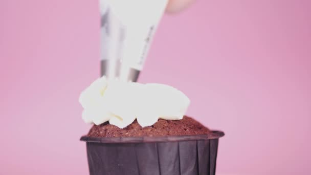 Aplicar crema blanca sobre un cupcake de chocolate negro sobre un fondo rosa. — Vídeos de Stock