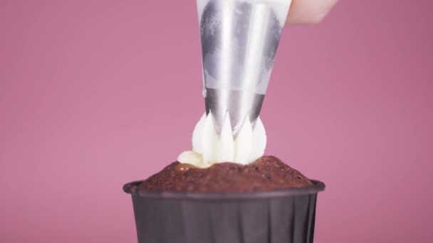 Применение белого крема на темно-шоколадный кекс кекс на розовом фоне. — стоковое видео