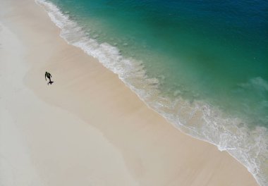 Yazın beyaz kumsalda yürüyen bir adamın üst görüntüsü. Deniz dalgaları berrak ve güzel renkli bir fotoğraf.