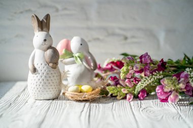 İki Paskalya porseleni tavşanı beyaz ahşap bir masanın üzerinde duruyor. Pençelerinde Paskalya yumurtaları var. Paskalya bayramında, parlak bahar çiçeklerinden oluşan bir kompozisyon