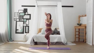 İnce, esnek, orta yaşlı, bej eşofmanlı siyah saçlı, beyaz bir kadın mor yoga minderinin üstünde asana taklidi yapıyor.