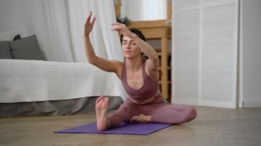 İnce, esnek, beyaz, orta yaşlı, siyah saçlı, bej eşofmanlı kadın Asana Ardha, Baddha, Padma, Paschimottanasana 'yı mor yoga minderinde, parlak yataklı bir pencerede,