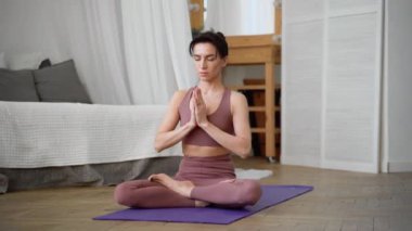 İnce, esnek, beyaz, orta yaşlı, siyah saçlı, bej eşofmanlı kadın mor yoga minderinin üzerinde asanas pranayamasını gevşetiyor.