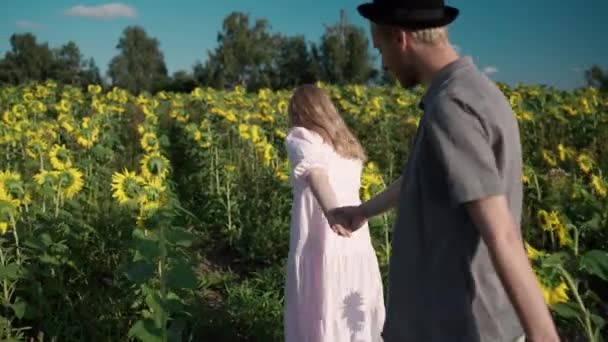 Joven chica rubia caucásica sonríe, lleva a chico en sombrero a través del campo de girasoles — Vídeos de Stock