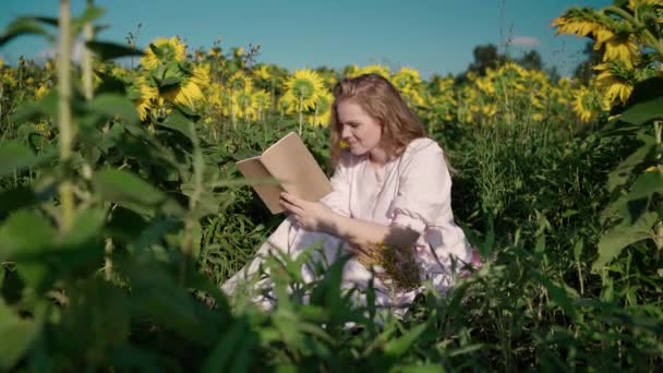 Seorang gadis muda berambut pirang di ladang bunga matahari membaca sebuah buku, melihat sekeliling, menunggu — Stok Video