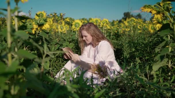 Uma jovem loira em um campo de girassol lê um livro, olha em volta, espera — Vídeo de Stock