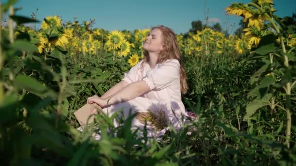 Uma jovem loira em um campo de girassol lê um livro, olha em volta, espera — Vídeo de Stock