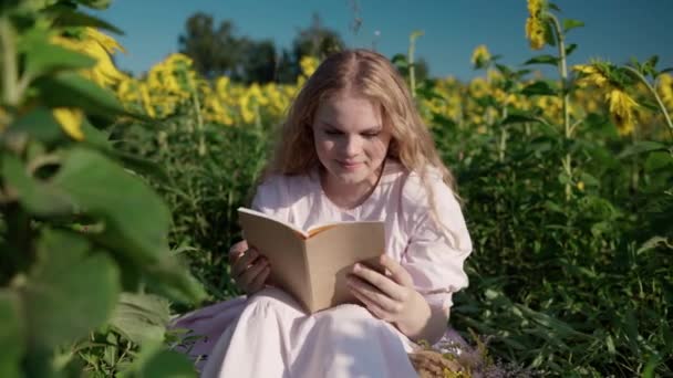 Ein junges blondes Mädchen in einem Sonnenblumenfeld liest ein Buch, schaut sich um, wartet — Stockvideo