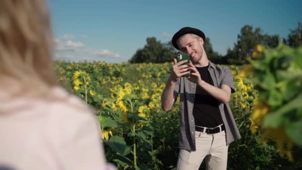 Chica rubia en un campo de girasol lee libro, posando en el teléfono a chico lindo — Vídeos de Stock