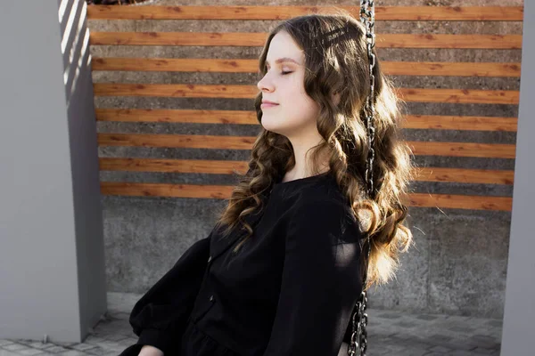 Portrait of a smiling young woman in black dress with closed eyes enjoying of bright sunlight sitting on wooden swing on street. Girl having fun outside. Urban style and fashion. Casual outfit — Stock Photo, Image