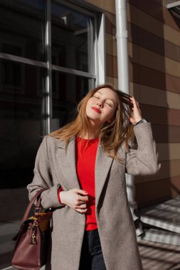 Palto giymiş gülümseyen genç bir kadının portresi saçlarına elle dokunuyor ve pencerenin önünde duran parlak güneş ışığının tadını çıkarıyor. Kız dışarıda eğleniyor. Şehir tarzı ve moda. Günlük kıyafet.