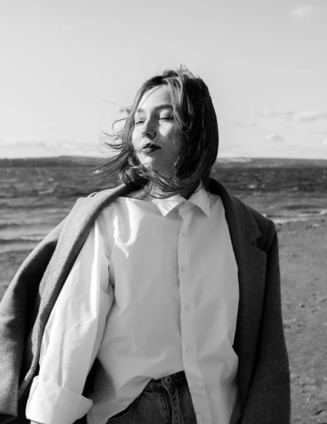 Portrait of a young woman in coat, white shirt and jeans with closed eyes enjoying of bright sunlight standing on beach in front of sea. Girl having fun outside. Urban style and fashion. Casual outfit — Stock Photo, Image