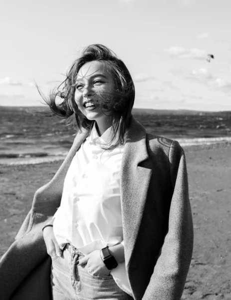 Portrait of a laughing young woman in coat, white shirt and jeans enjoying of bright sunlight standing on beach in front of sea. Girl having fun outside. Urban style and fashion. Casual outfit — Stock Photo, Image