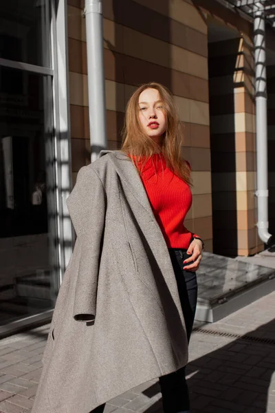 Portrait of a young woman taking off coat, looking at camera and standing on street in front of window and brown wall. Caucasian girl having fun outside. Urban style and fashion. Casual outfit — Stock Photo, Image