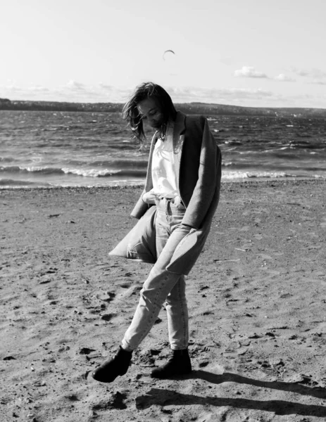 Retrato de una joven con abrigo, camisa blanca y jeans disfrutando de la luz del sol en la playa frente al mar. Chica divirtiéndose afuera. Estilo urbano y moda. Traje casual —  Fotos de Stock