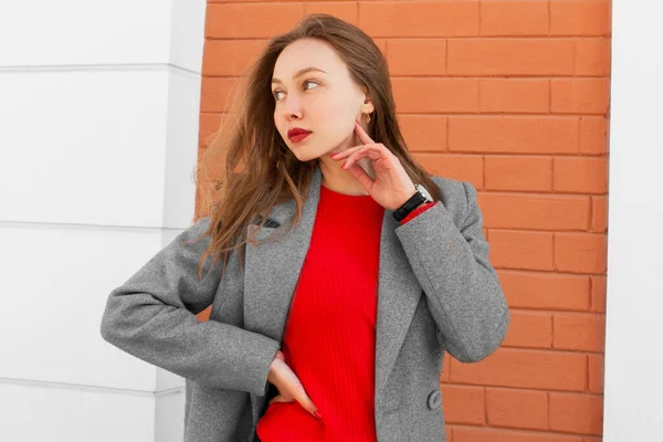 Καστανά μαλλιά νεαρή γυναίκα σε παλτό, κόκκινο πουκάμισο με ρολόγια στο χέρι, στέκεται στο δρόμο και κοιτάζοντας την αριστερή πλευρά μπροστά από το κόκκινο τοίχο τούβλο. Λευκή κοπέλα. Αστικό στυλ και μόδα. Απλό ντύσιμο — Φωτογραφία Αρχείου