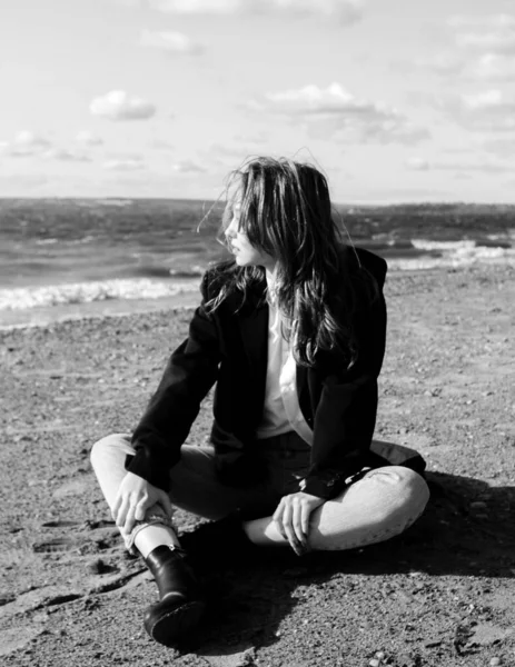 Portrait of a laughing young woman in black jacket, white shirt and jeans enjoying of sunlight sitting on beach sand in front of sea. Girl having fun outside. Urban style and fashion. Casual outfit