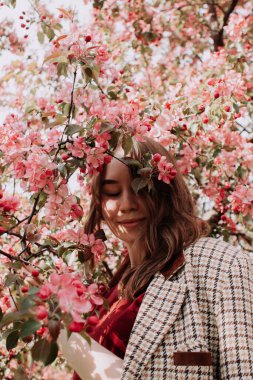 Ceketli gülümseyen genç bir kadın çiçek açan elma veya kiraz ağacının pembe çiçekleri arasında tek başına parlak güneş ışığının tadını çıkarıyor. Çiçekli kız. Yazlık kıyafet. Bahar günü.