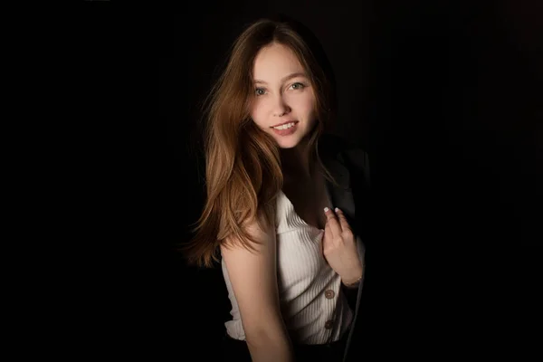 Young woman in jacket and white t-shirt standing alone in dark room and looking at camera, black background. Studio shot in low key. Casual portrait of girl with natural makeup and clean skin.