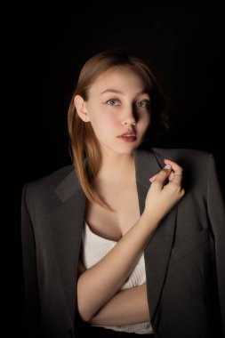 Ceketli ve beyaz tişörtlü genç bir kadın karanlık odada dikiliyor ve siyah arka planda kameraya bakıyor. Stüdyo görüntüsü sade. Doğal makyajlı ve temiz tenli bir kızın sıradan portresi..