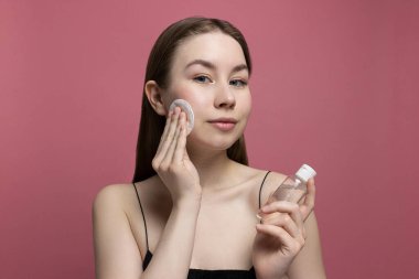 Gülümseyen genç bir kadın, elinde pembe arka planda bir şişe mikiler suyu tutarak pamuk pedi ile makyajını temizliyor. Temizlikçi kız. Sağlıklı tedavi ve kozmetoloji ürünleri. Güzellik rutini, cilt bakımı