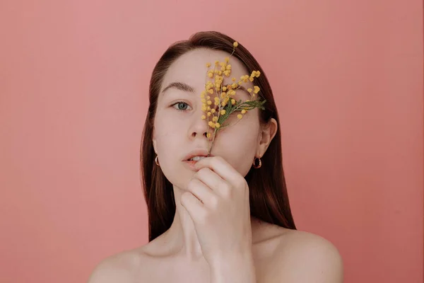 Smiling young woman with naked shoulders and no makeup holding branch of yellow mimosa flower on pink background studio shot. Smiling girl with brown hair. Spring, summer concept. Natural beauty