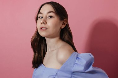 Mavi gömlekli, çıplak omuzlu, doğal makyajlı, kıvırcık saçlı pembe arka planda kameraya bakan genç bir kadın, gölge. İnci küpeli günlük giysili beyaz kız. Stüdyo fotoğrafı..