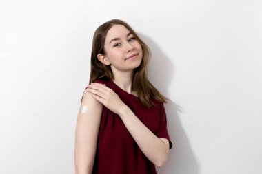 Tişörtünde Coronavirus aşısından sonra omuzlarında alçı olan gülümseyen genç bir kadın, beyaz arka plan, fotokopi alanı. Aşı, aşı, grip önleme konsepti. Covid-19 aşısı alınıyor