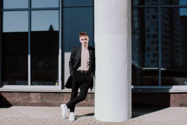 Siyah ceketli ve pantolonlu yakışıklı bir genç pencerenin önünde parlak bir güneş ışığına yaslanmış beyaz bir sütuna yaslanıyor. Sokak modası. Kameraya bakan akıllı, sıradan tarzda bir adam.