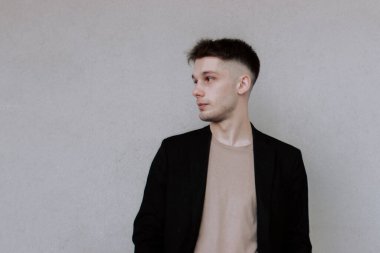 Siyah ceketli, kahverengi saçlı yakışıklı genç adam gri beton duvarın önünde tek başına duruyor, sola bakıyor, boşluğu kopyalıyor, yandan bakıyor. Şehir stye ve sokak modası. Şık giyinmiş bir adam.