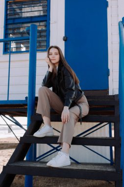 Siyah deri ceketli gülümseyen genç bir kadın mavi kapının önünde gözetleme kulesinin merdivenlerinde oturuyor. Dışarıda eğleniyoruz. Şehir tarzı ve sokak modası. Sıradan kıyafetli bir kız kameraya bakıyor.