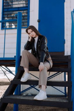 Siyah deri ceketli gülümseyen genç bir kadın mavi kapının önünde gözetleme kulesinin merdivenlerinde oturuyor. Dışarıda eğleniyoruz. Şehir tarzı ve sokak modası. Sıradan kıyafetli bir kız kameraya bakıyor.