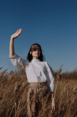 Beyaz bluzlu genç bir kadın gökyüzünün önündeki kuru otların arasında duruyor. Parlak güneş ışığıyla yüzünü kaplıyor. Tarz ve moda. Kapalı gözlü, sıradan giysili bir kız. Altın saat