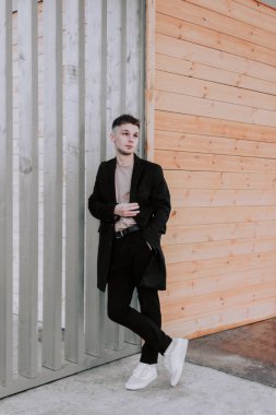 Siyah ceketli, kahverengi saçlı, beyaz ayakkabılı, gri ve tahta duvarın önünde tek başına sağa bakan yakışıklı bir genç. Şehir stye ve sokak modası. Şık giyinmiş bir adam.