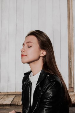 Siyah deri ceketli genç bir kadın beyaz duvarın önündeki tahta merdivenlerde oturuyor. Profil görünümü portresi. Güneş ışığının tadını çıkarıyorum. Şehir tarzı ve sokak modası. Kapalı gözlü, günlük kıyafetli bir kız..