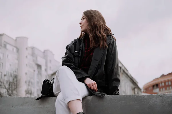 Leende ung kvinna i svart skinnjacka och vita jeans sitter på en betong och tittar åt vänster. Urban stil och street mode. Flicka med lockig frisyr i avslappnad outfit. Profilbild — Stockfoto