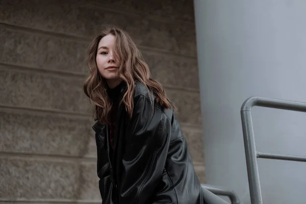 Smilende ung kvinde i sort læderjakke stående foran grå væg, lænet på søjlen, ser på kameraet. Urban stil og street fashion. Pige med krøllet frisure i afslappet tøj - Stock-foto